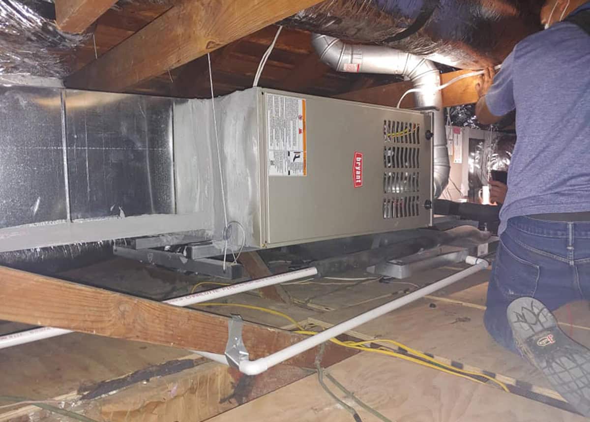 HVAC technician servicing a Bryant gas furnace installed in a DFW attic, showing ductwork, drain lines, and heater components during a furnace tune-up by Bolton Heating & Air.