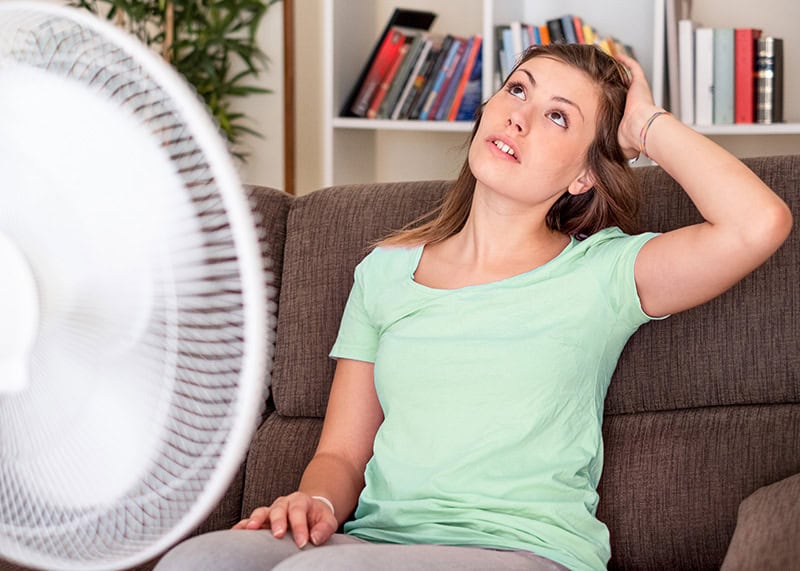 Woman feeling hot and frustrated sitting on couch with fan because AC is running but not cooling her Forney Texas home